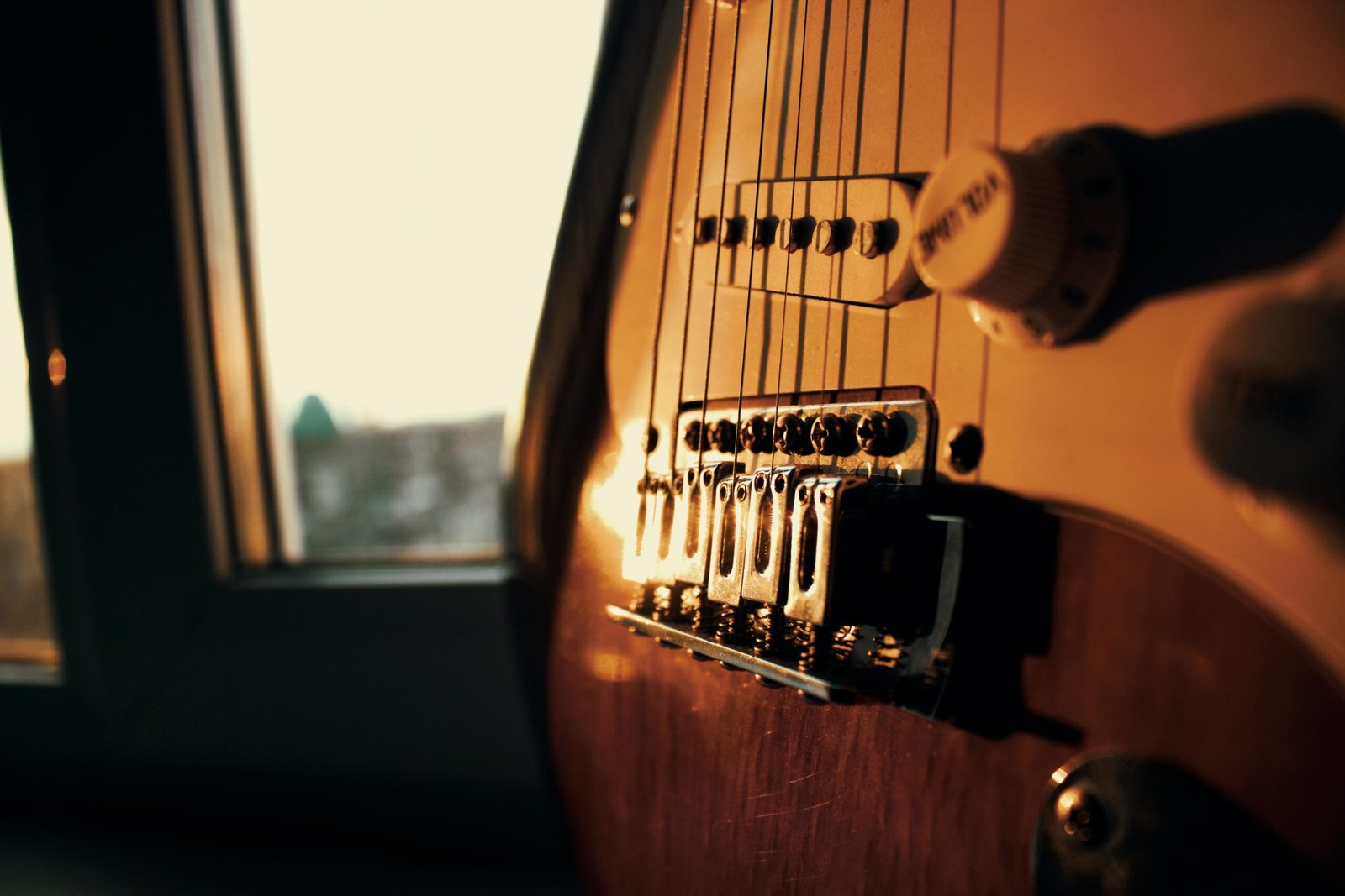 Ambient close-up of an electric guitar's bridge illuminated by sunset light through a window.
